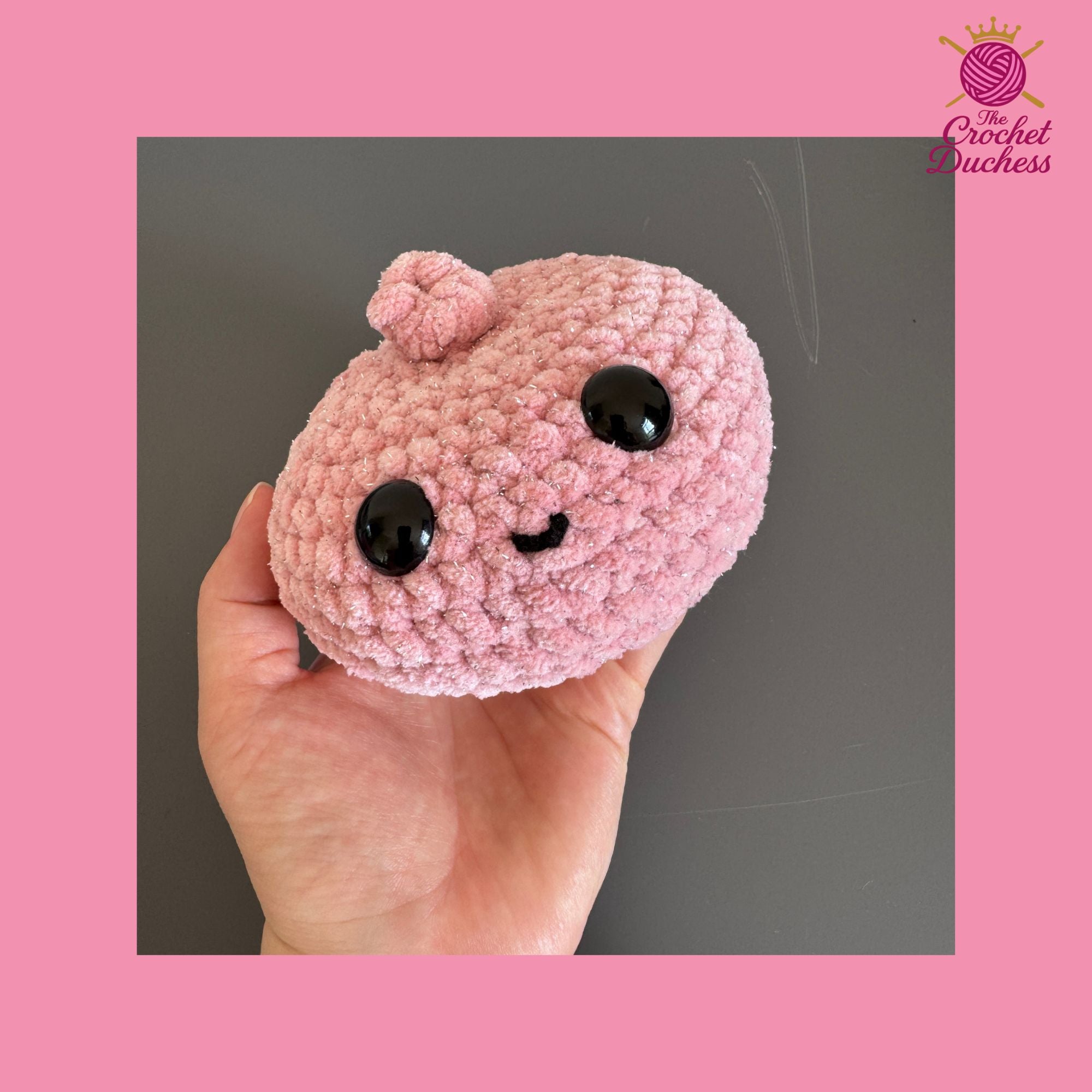 A pink crocheted plush dumpling with a cute face and black eyes, held in a person's hand against a dark gray background, framed by a pink border with 'The Crochet Duchess' logo in the top right.