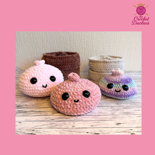 Four cute crocheted plush toys shaped like round characters with large black eyes and simple stitched smiles, displayed on a wooden surface with three matching crocheted storage baskets behind them. The main foreground toy is light pink, with other variants in brown, beige, and purple.