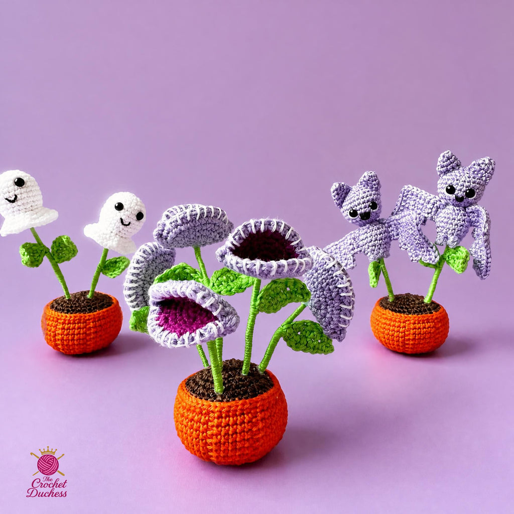 Four crocheted amigurumi plant toys arranged on a lavender background. The set includes two white ghost flowers, two lavender-and-white flower pots with purple flowers, and one orange carrot-shaped pot with a purple bat in a flower pot.