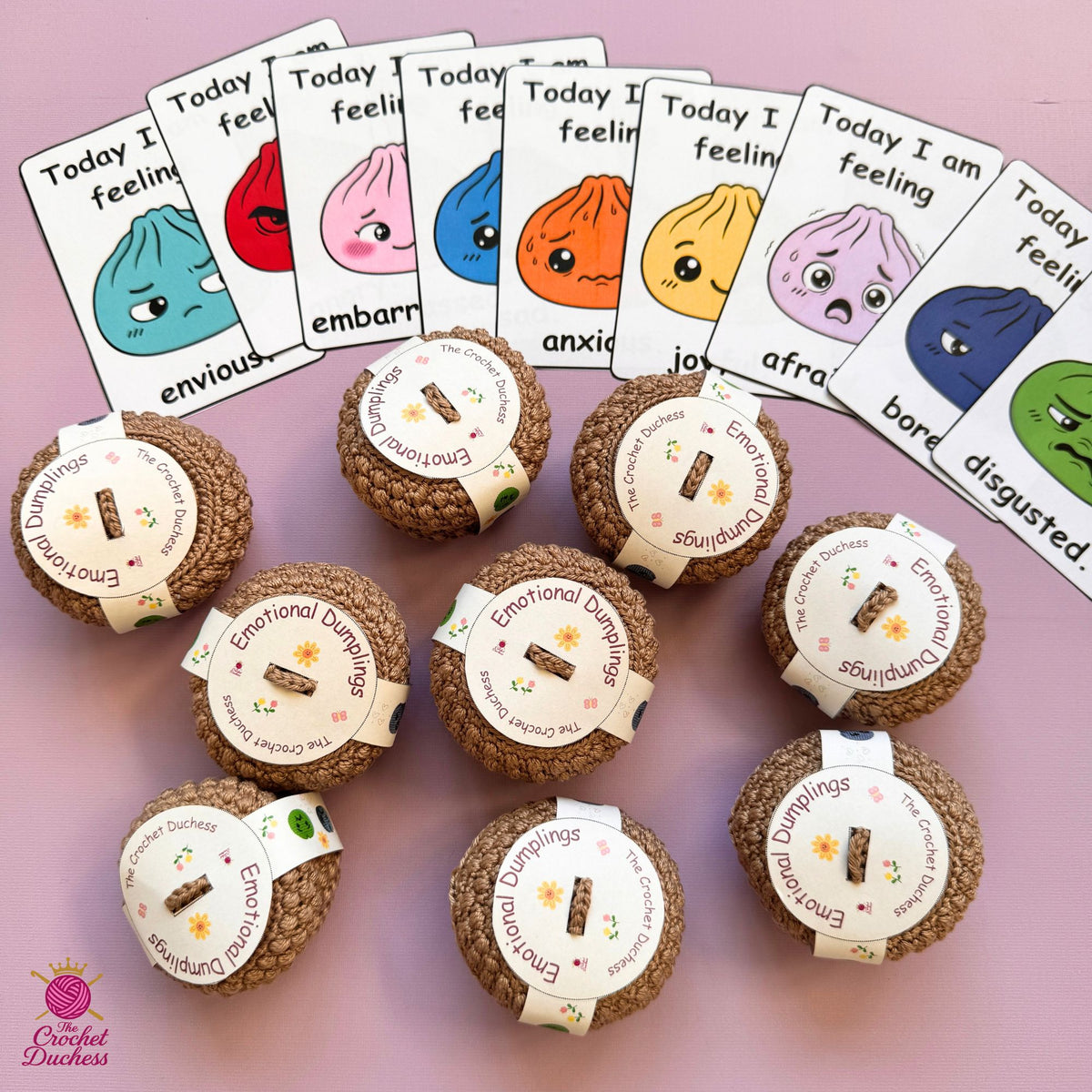 A set of brown textured dumpling-shaped items wrapped with white paper labels reading 'Emotional Dumplings' arranged on a pink background, with matching emotion cards fanned out behind them. A 'Crochet Duchess' logo appears in the lower left corner.
