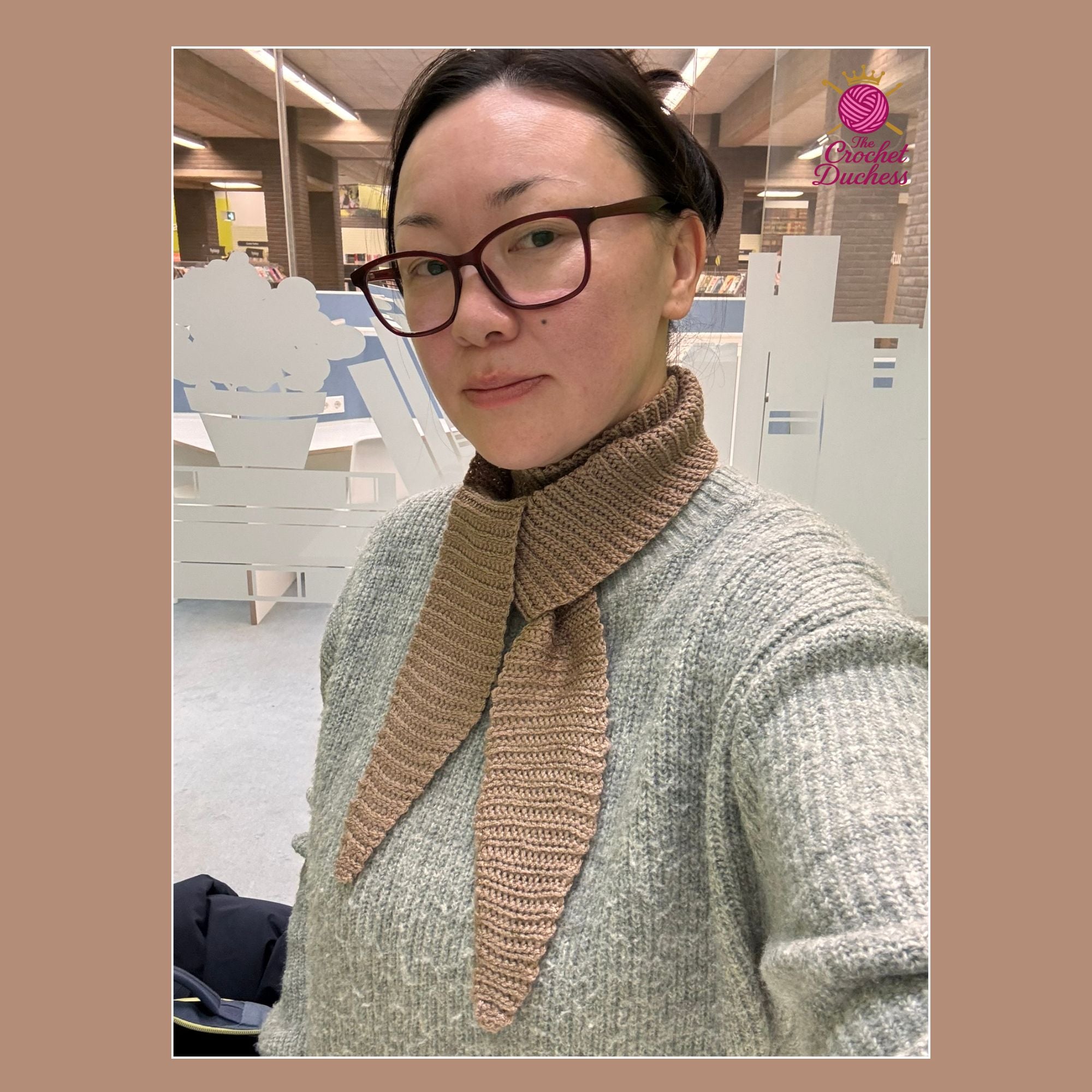 A woman wearing a chunky crochet brown scarf with a wide collar over a gray knit sweater, photographed in a modern indoor setting with frosted glass partitions. A pink 'The Crochet Duchess' logo is visible in the top right corner.