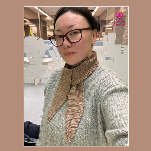 A woman wearing a chunky crochet brown scarf with a wide collar over a gray knit sweater, photographed in a modern indoor setting with frosted glass partitions. A pink 'The Crochet Duchess' logo is visible in the top right corner.