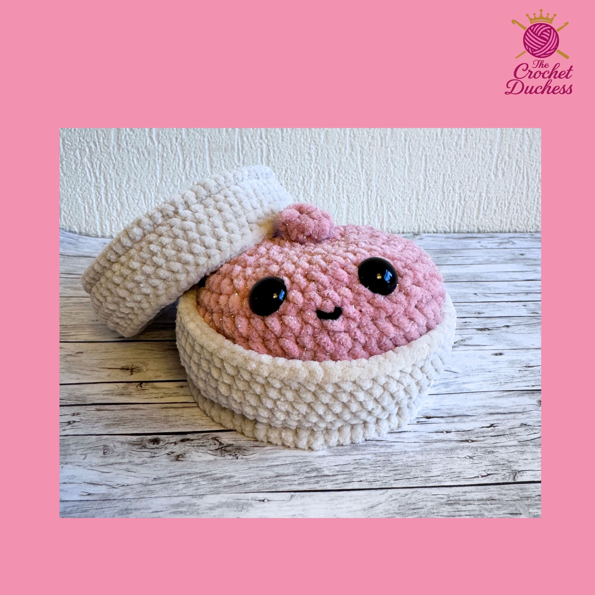 A crocheted amigurumi-style dumpling in pink with large black eyes and a small stitched smile, sitting inside a cream-colored crocheted cup or basket. A matching cream crocheted lid or cover is placed behind it on a rustic wood surface against a textured wall.