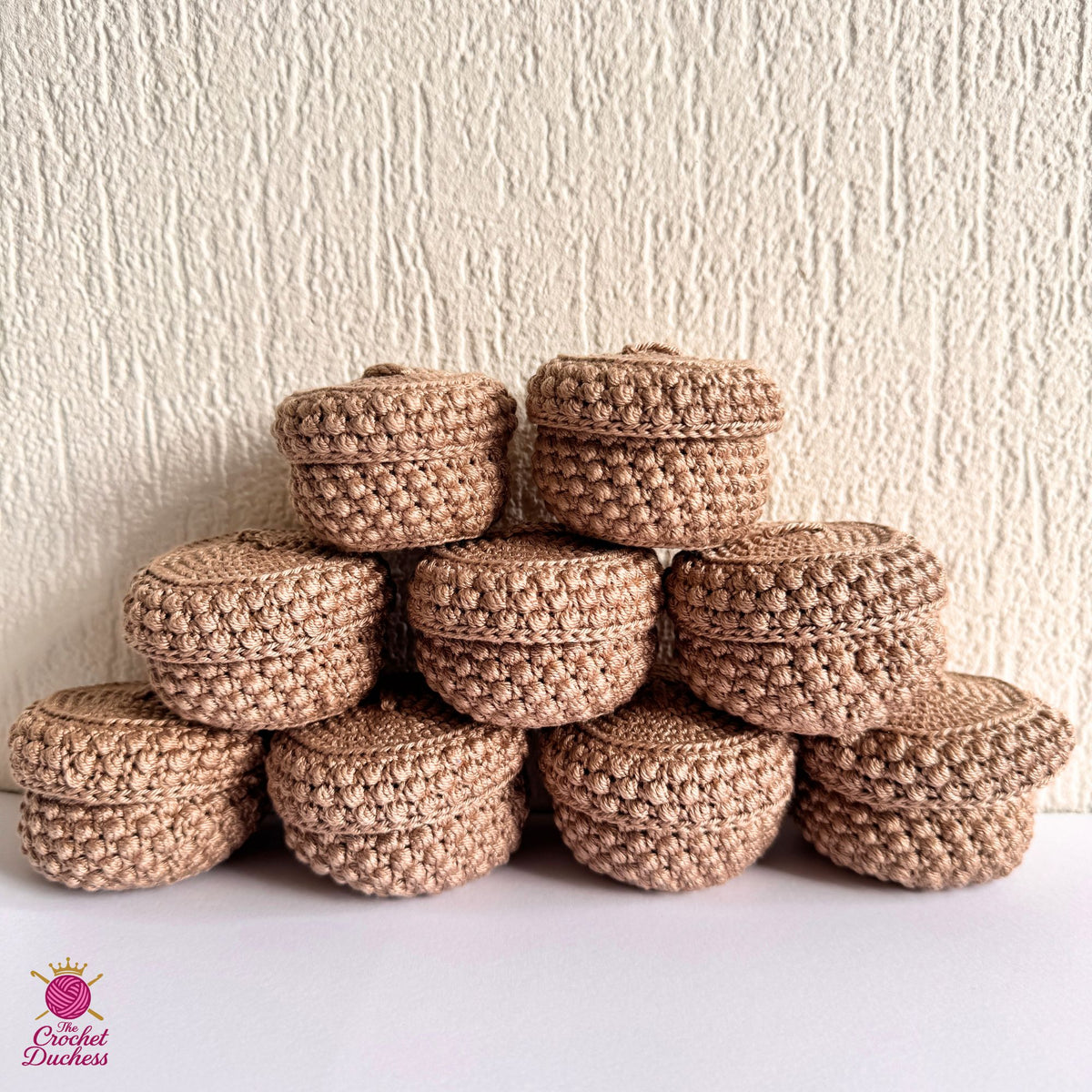 A stack of nine small round crocheted steamers with lids, made from a cotton beige yarn, arranged on a white surface against a textured beige wall. A 'Crochet Duchess' logo is visible in the bottom-left corner.