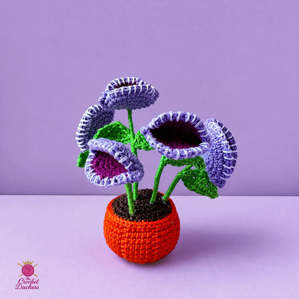 A crocheted Venus Flytrap in a small orange pumpkin pot, featuring five purple and lavender flowers with green stems and leaves, set against a solid lavender background.