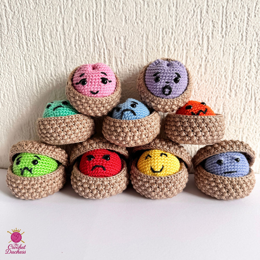 A group of nine crocheted amigurumi dumplings characters in various colors and expressions, arranged in two rows on a white surface against a textured beige wall. A 'Crochet Duchess' logo is visible in the bottom-left corner.
