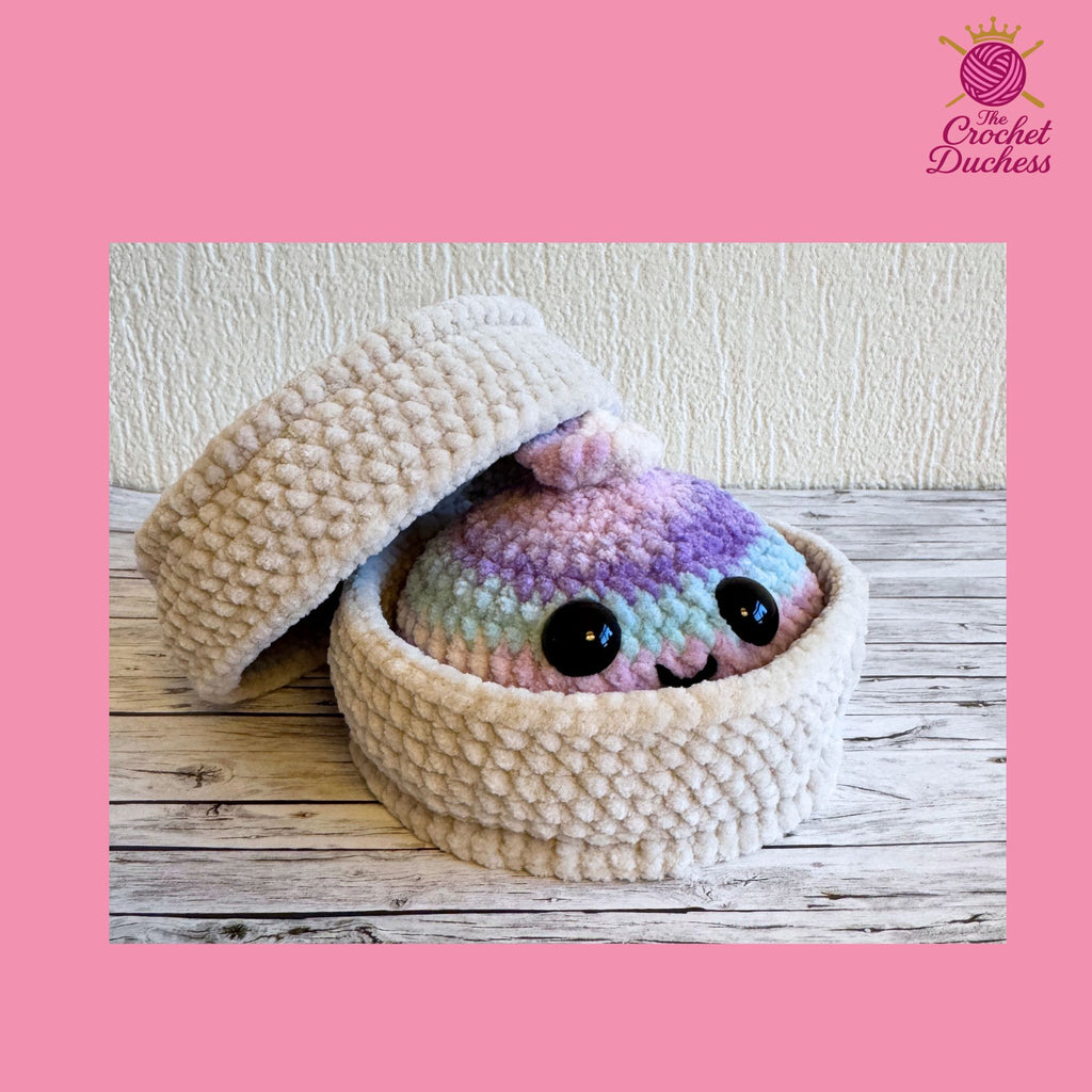 A handmade crochet amigurumi dumpling peeking out from a textured cream-colored basket, set on a rustic wooden surface with a textured wall background. The character has pastel pink, purple, and blue yarn colors with black eyes.