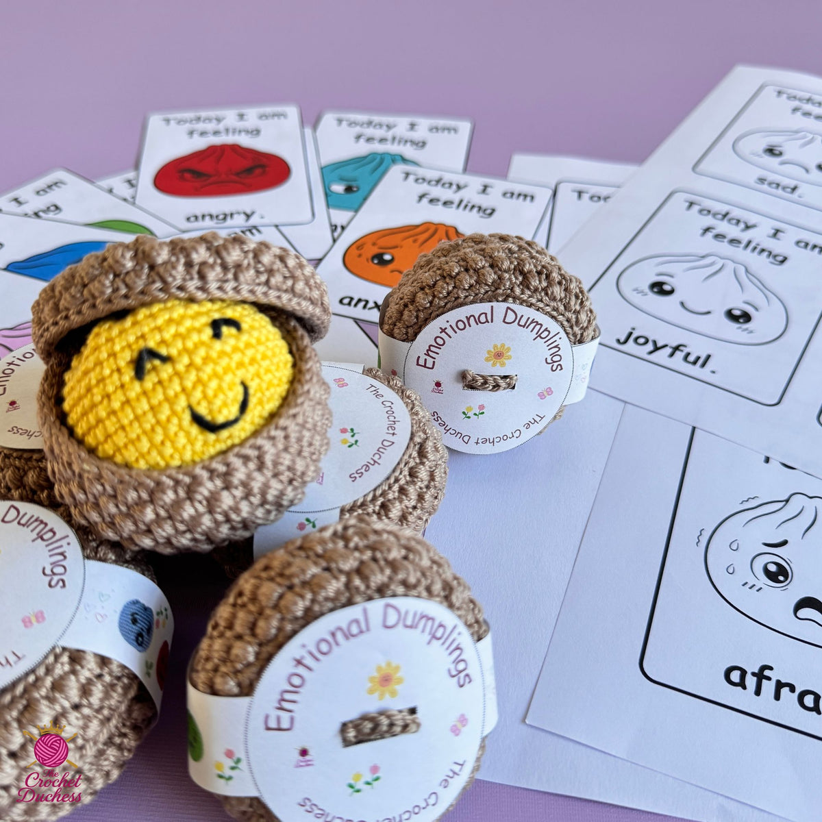 A close-up of handmade crocheted emotional dumpling toys arranged on a lavender surface, with printed emotion cards scattered behind them. The main product is a brown yarn dumpling with a yellow crocheted face peeking out, and similar dumplings are visible around it.