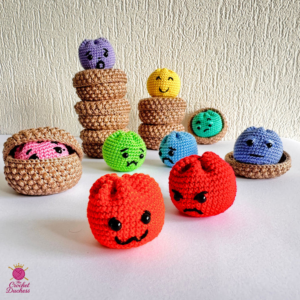A collection of handmade crocheted amigurumi dumplings, including two red angry faces in the foreground, a pink face inside a basket on the left, and several colorful round characters in different colored baskets and stacks, set against a textured off-white wall and white surface.