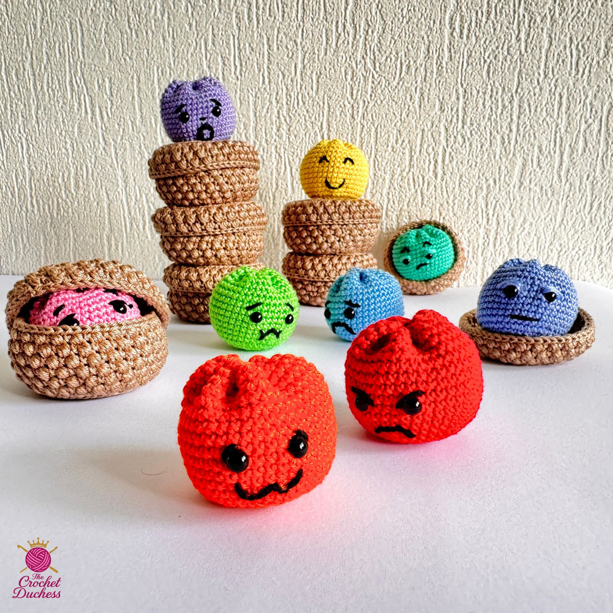 A collection of handmade crocheted amigurumi dumplings, including two red angry faces in the foreground, a pink face inside a basket on the left, and several colorful round characters in different colored baskets and stacks, set against a textured off-white wall and white surface.